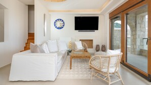 Una sala de estar moderna con un sofá blanco, una mesa de centro de madera y un sillón de mimbre. Un televisor de pantalla plana está montado en la pared sobre una chimenea.