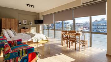 Un dormitorio moderno con una cama grande, un sofá colorido, una mesita pequeña y vistas a la ciudad a través de amplias ventanas.