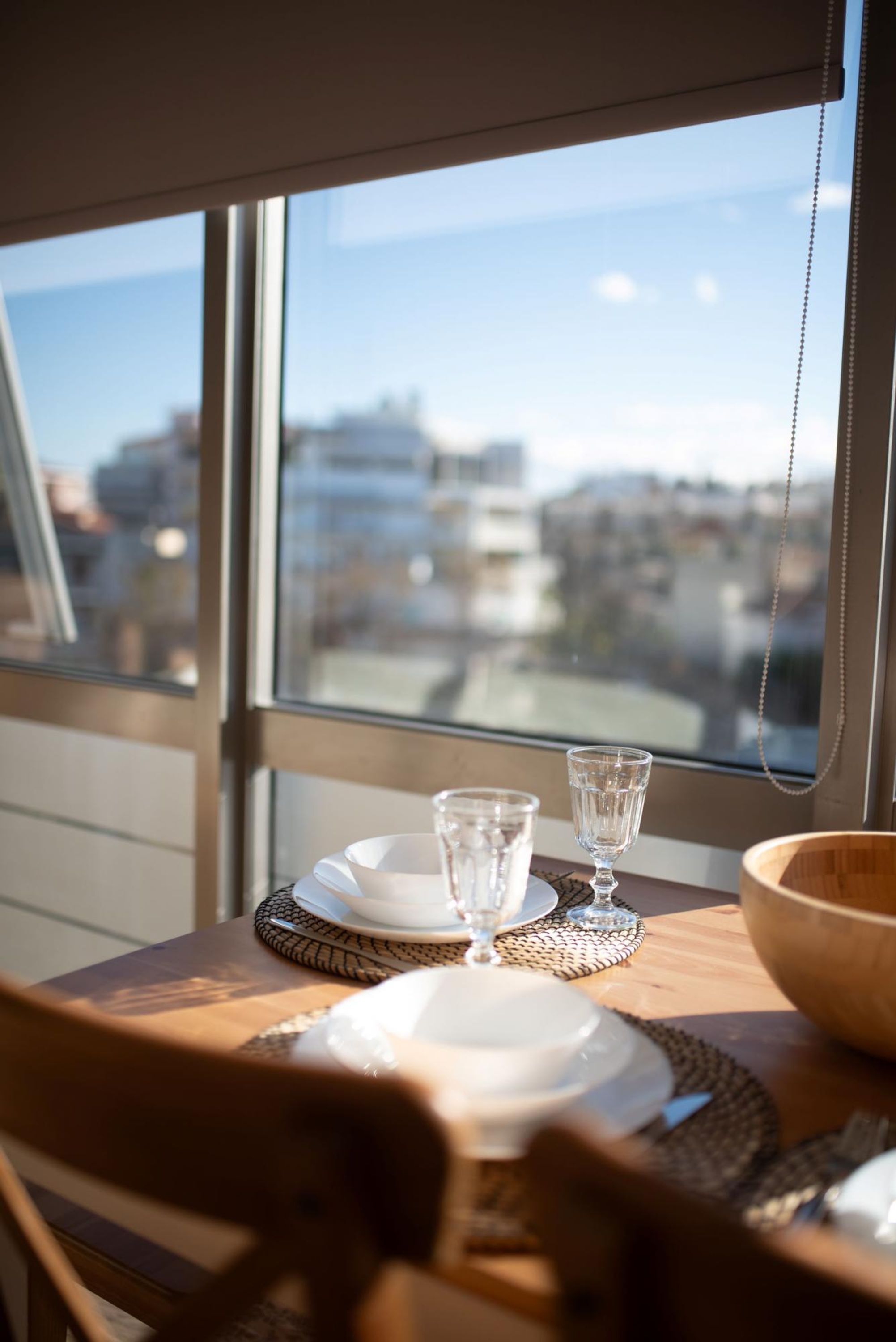 Una mesa de comedor con platos, vasos y un bol, frente a una ventana con vistas a la ciudad.