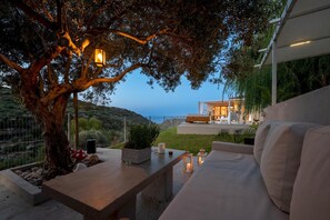 A modern outdoor seating area with a view of the sea, a tree, and a building with a white facade. - Canyon 1 Elegant villa (Rethymno)