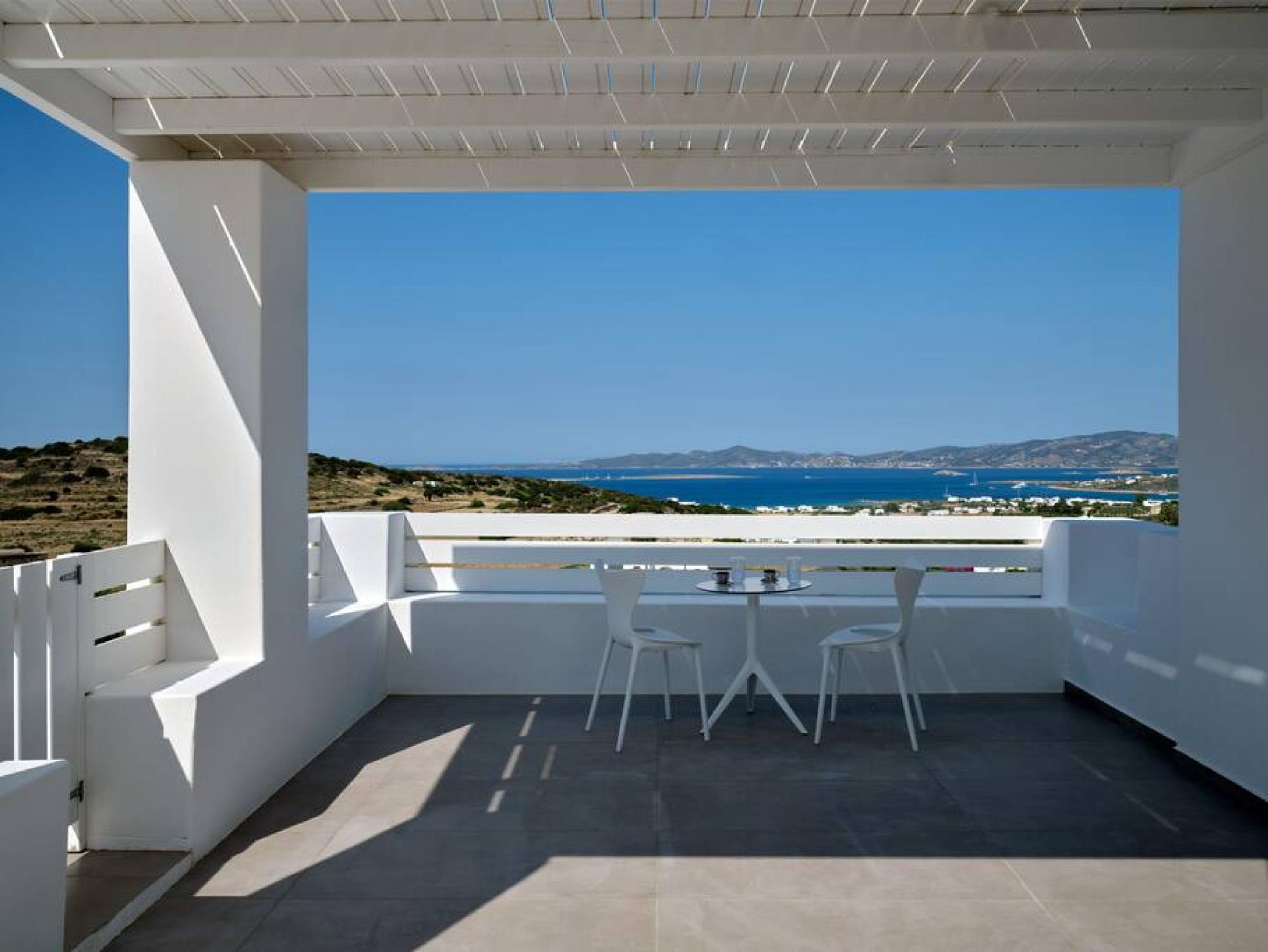 A balcony with a table and chairs, overlooking a coastal landscape with a clear sky.