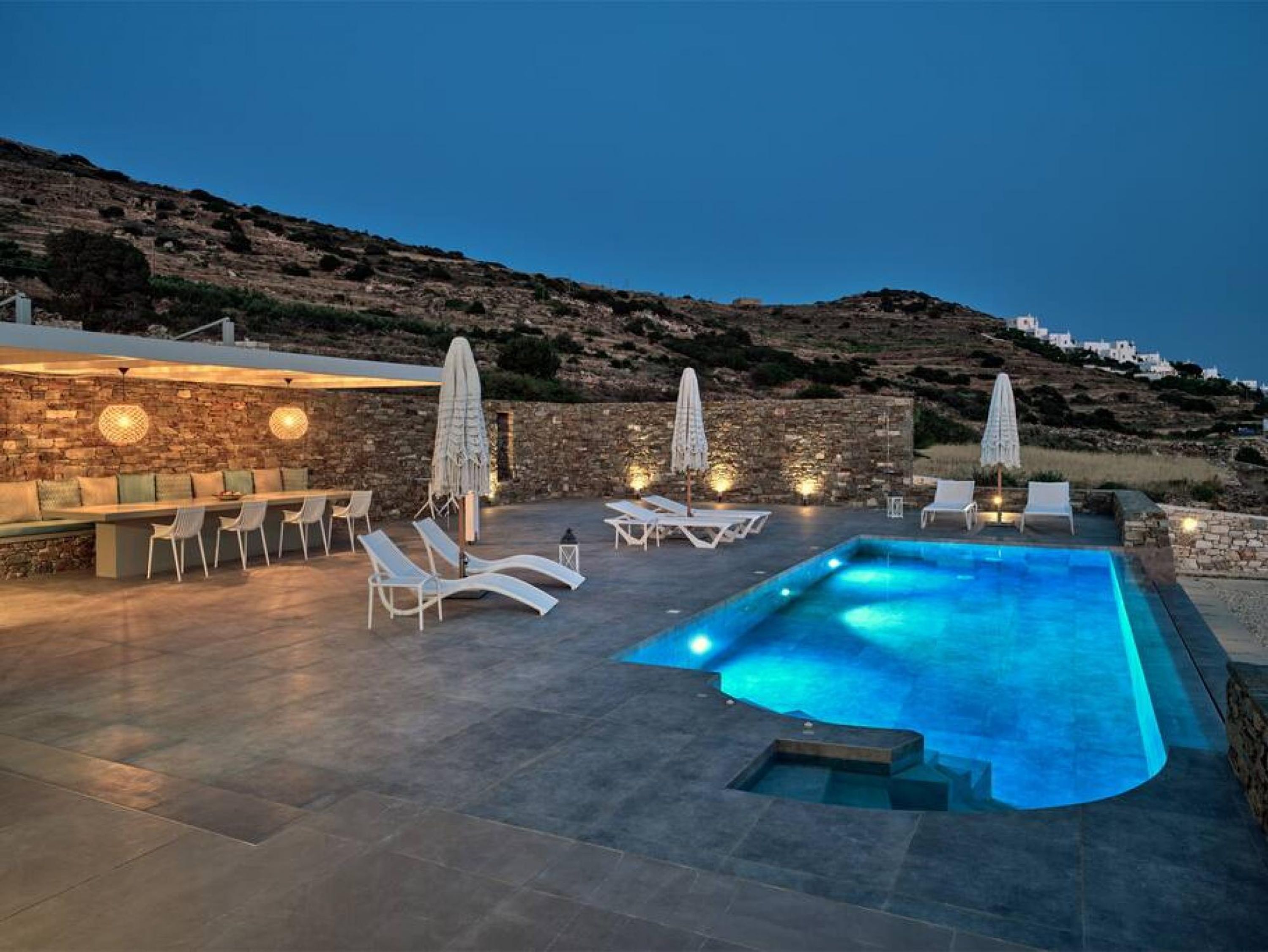 Un espace piscine doté de chaises longues et d’un bar, entouré de murs en pierre et offrant une vue sur une colline.