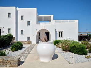 A modern white building with a central entrance, a stone pathway, and a large white planter with plants. - La Vitalite Luxury Villa Mer in Paros (Aliki)