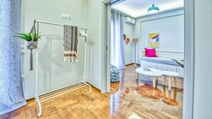 Una sala moderna con un sofá, una mesa de centro y una pequeña mesa de comedor. Hay una planta en un estante, un perchero y un aplique de pared.