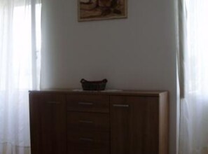 Ein Zimmer mit einem Holzkabinett, einem gerahmten Bild an der Wand und transparenten Vorhängen.