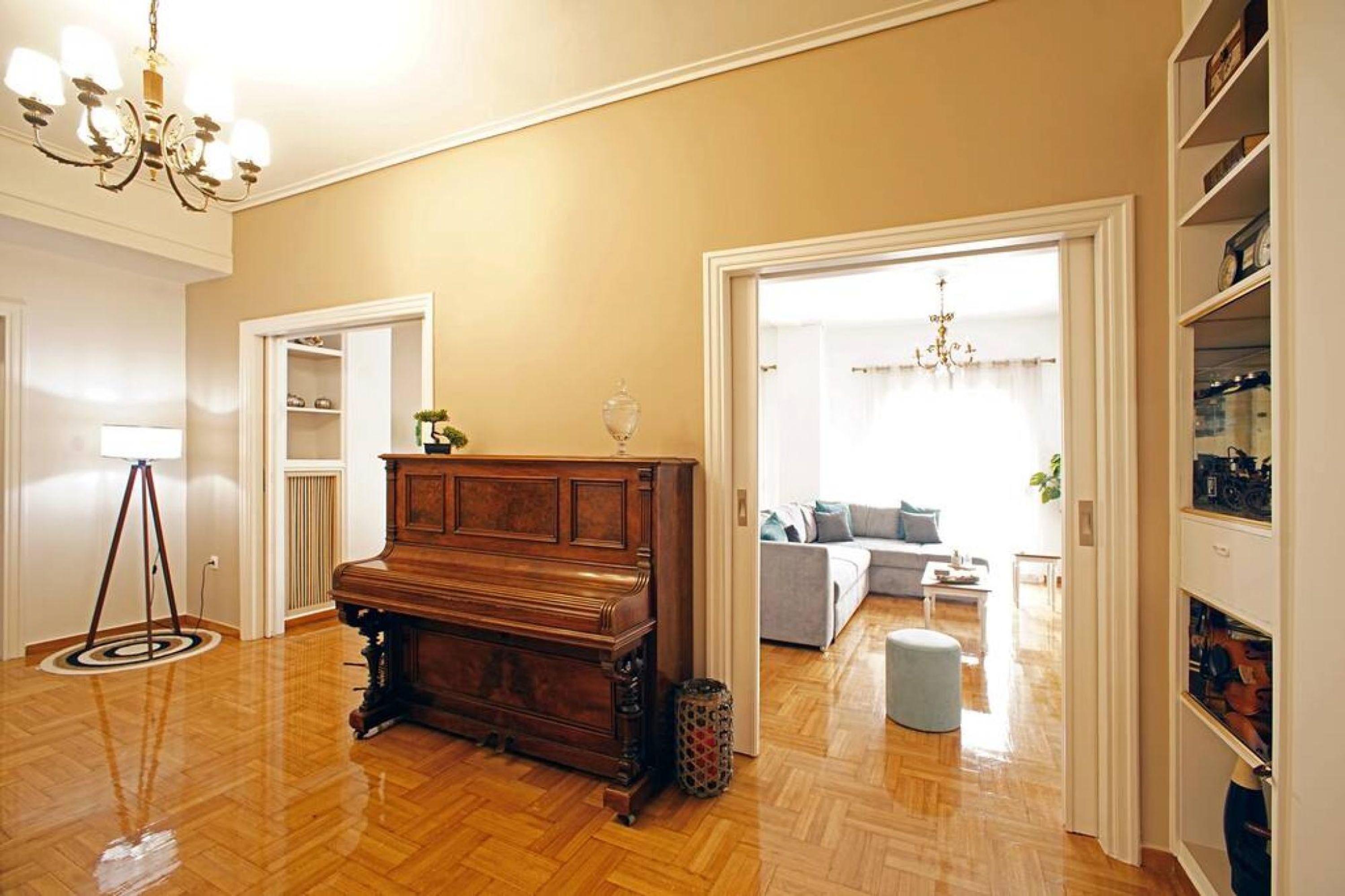 A spacious living room with a wooden piano, a chandelier, and a bookshelf.