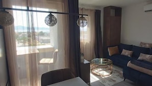 A living room with a sofa, a round coffee table, and a chair. There is a window with sheer curtains and a view of the outside.