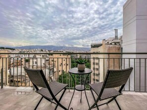 Ein Balkon mit zwei Korbstühlen und einem kleinen Tisch, mit Blick auf eine Stadtlandschaft mit Gebäuden und Bergen in der Ferne.