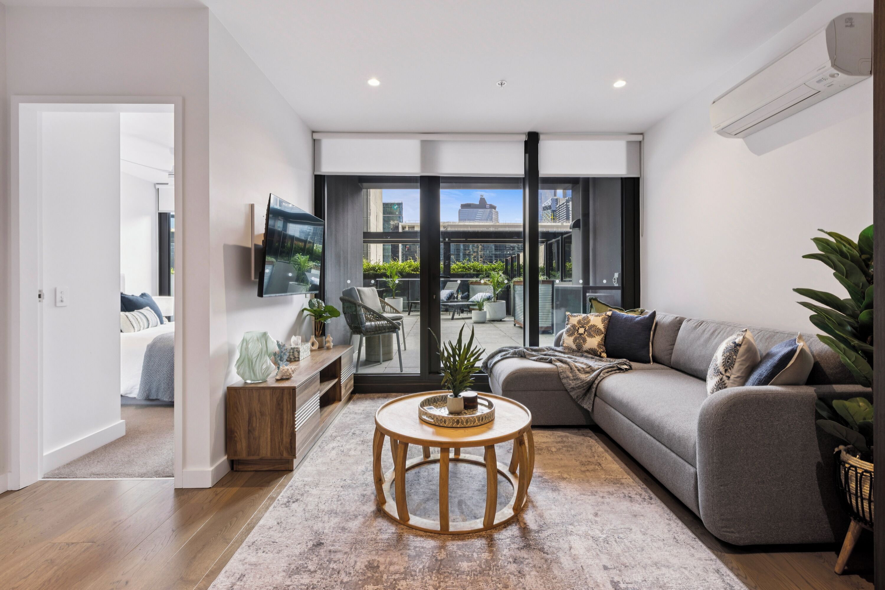 A modern living room with a grey sofa, a round wooden coffee table, and a large window offering a city view.
