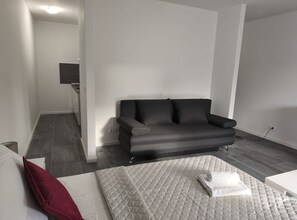 Un salon moderne avec un canapé gris, un lit blanc avec un coussin rouge et un matelas blanc matelassé.