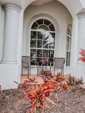A covered patio with two chairs, a table, and a large arched window overlooking a garden with red plants. - Beautiful home near Englewood Beach (Rotonda West)
