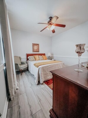 Una camera d'albergo con un letto, una sedia, una cassettiera in legno e una ventola a soffitto.