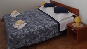 Siististi pedattu sänky, jossa on kukallinen vuodevaatesetti, kaksi tyynyä ja siististi taitetut pyyhkeet. Sängyn vieressä on yöpöytä, jossa on lamppu, ja lattialla on pieni matto.