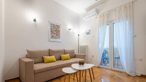 A living room with a beige sofa, a small white table, wooden flooring, a radiator, a picture on the wall, and a balcony with white curtains. - Roubnb Acropolis Museum 1BD Apt (Athina)