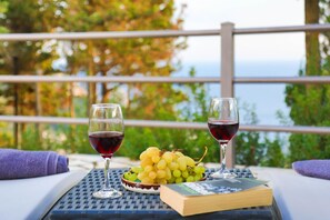 Ein Tisch mit zwei Weingläsern, einer Weintraubentafel und einem Buch auf einem Balkon mit Geländer und Grünpflanzen im Hintergrund.