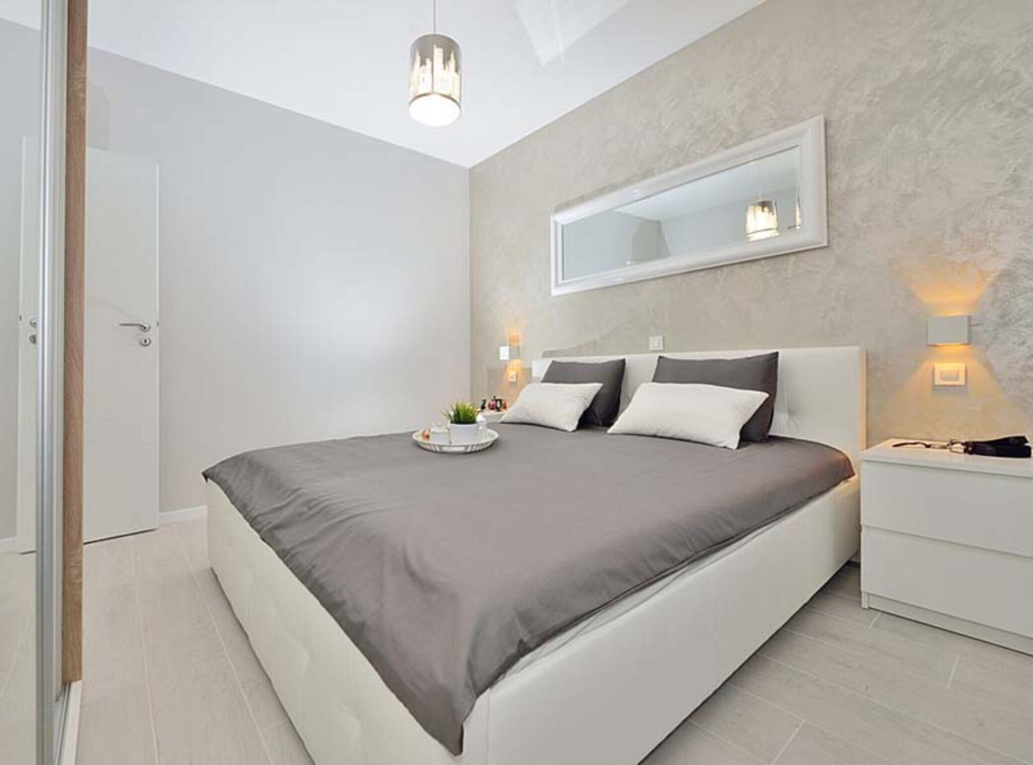 A modern bedroom with a large bed, bedside tables, a mirror, and a ceiling light.
