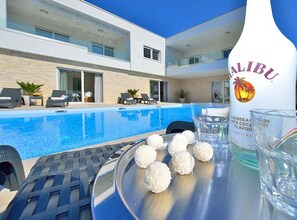 Uma área moderna à beira da piscina, com uma garrafa de rum Malibu, um tabuleiro com bolas de coco e espreguiçadeiras.