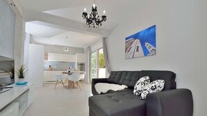 Un salon moderne avec un canapé foncé, un coussin blanc et moelleux, et un coussin noir et blanc à motifs. On y trouve également un coin repas avec une table et des chaises blanches, et une cuisine en arrière-plan.