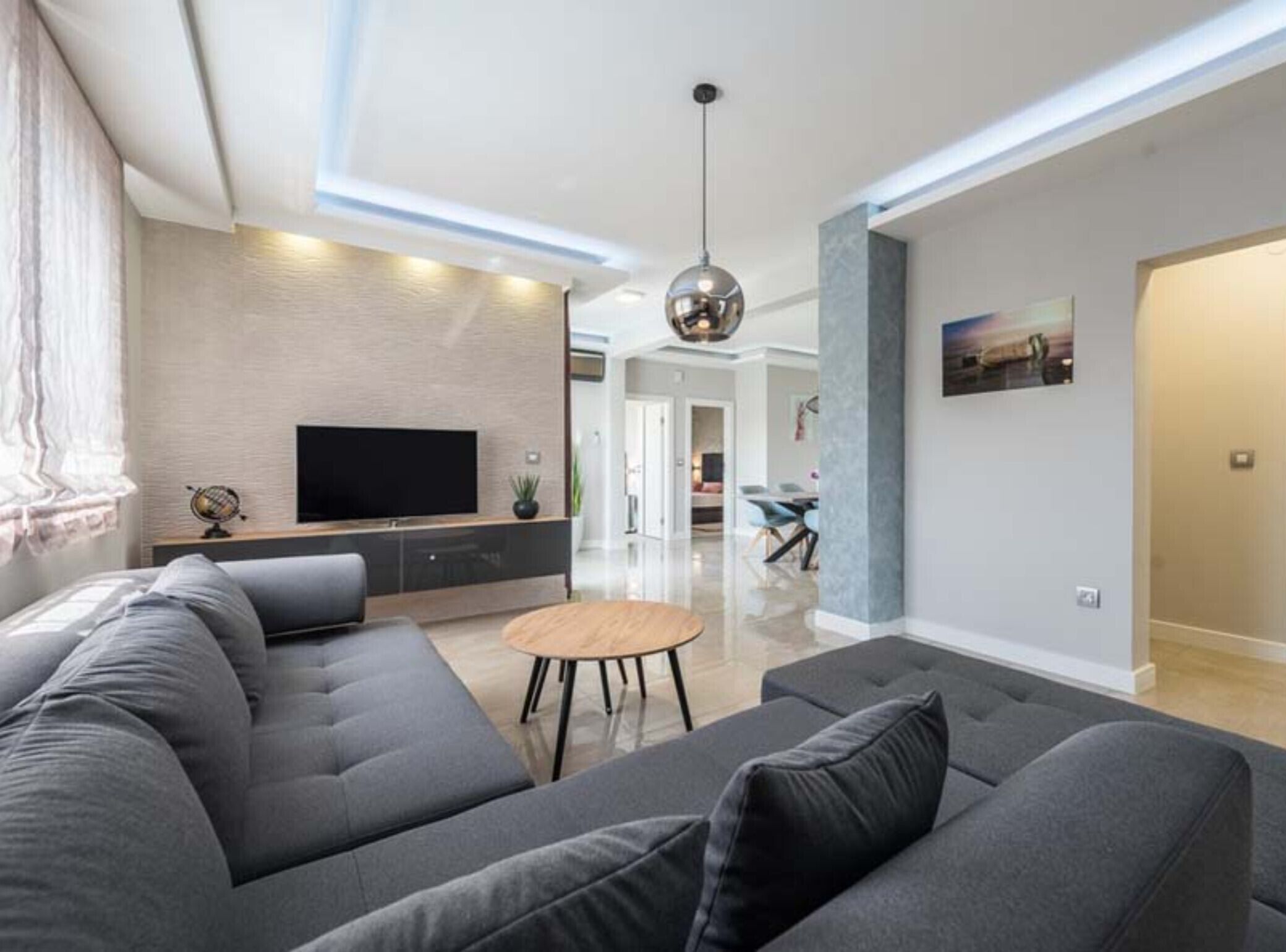 Ein modernes Wohnzimmer mit einem großen grauen Sofa, einem kleinen Holztisch und einem Flachbildfernseher, der an einer strukturierten Wand angebracht ist.