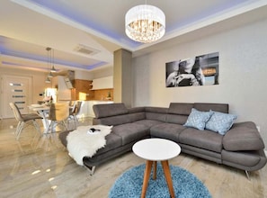 A modern living room with a sectional sofa, a white round table, a chandelier, and a kitchen in the background. - Villa Marija Exclusive Stay - Two Bedroom Apartment with Terrace 4 (Sukosan)