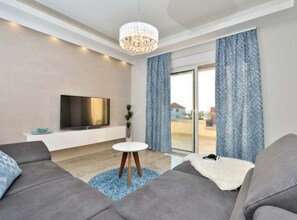 Ein modernes Wohnzimmer mit Flachbildfernseher, grauen Sofas, einem kleinen weißen Tisch und einer Chandelüster.