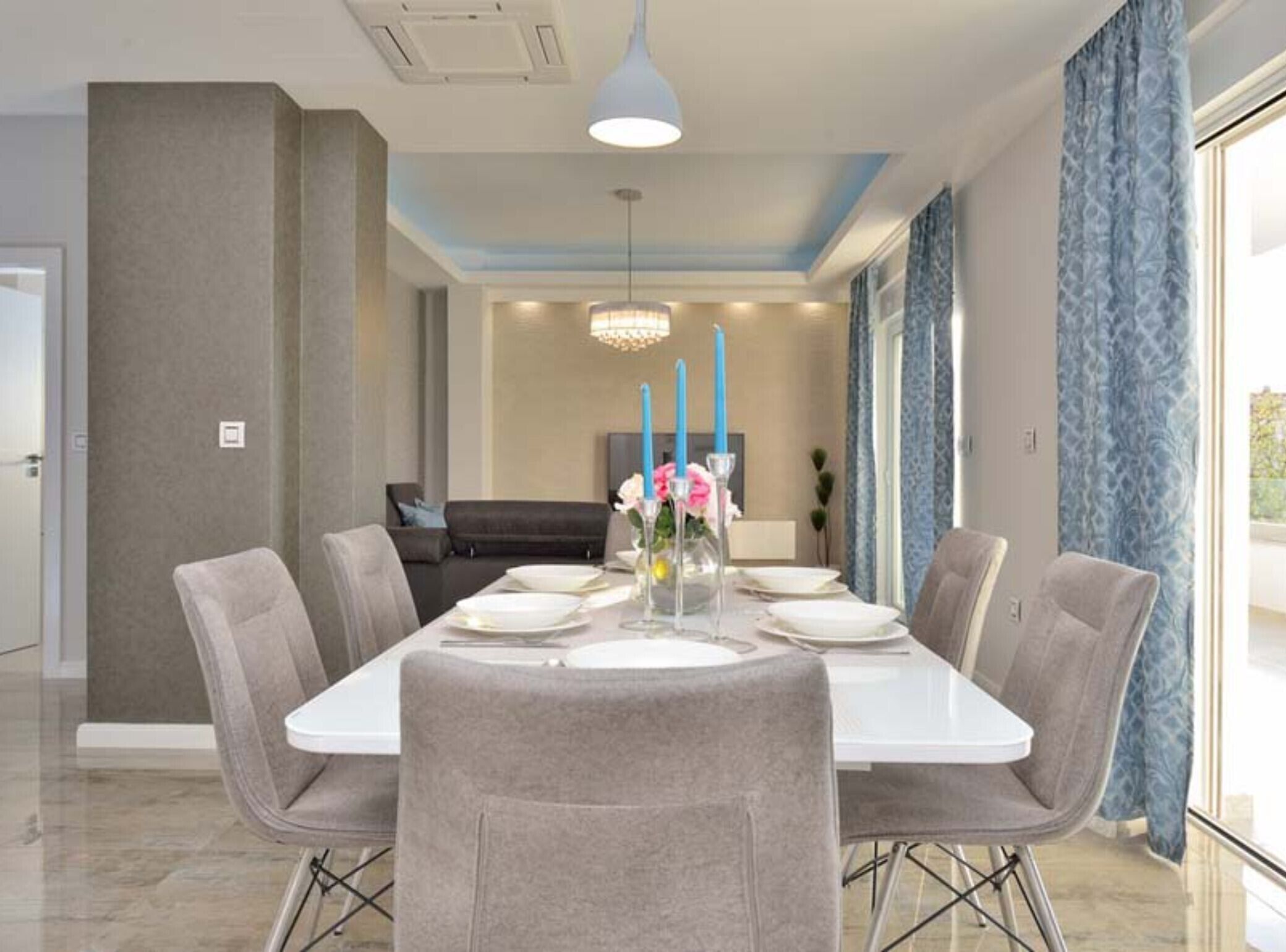 Ein modernes Esszimmer mit einem rechteckigen Tisch, gedeckt für sechs Personen, umgeben von sechs grauen Polsterstühlen. Eine Kristallleuchter hängt von der Decke, und im Hintergrund ist ein großes Fenster mit blauen Vorhängen zu sehen.