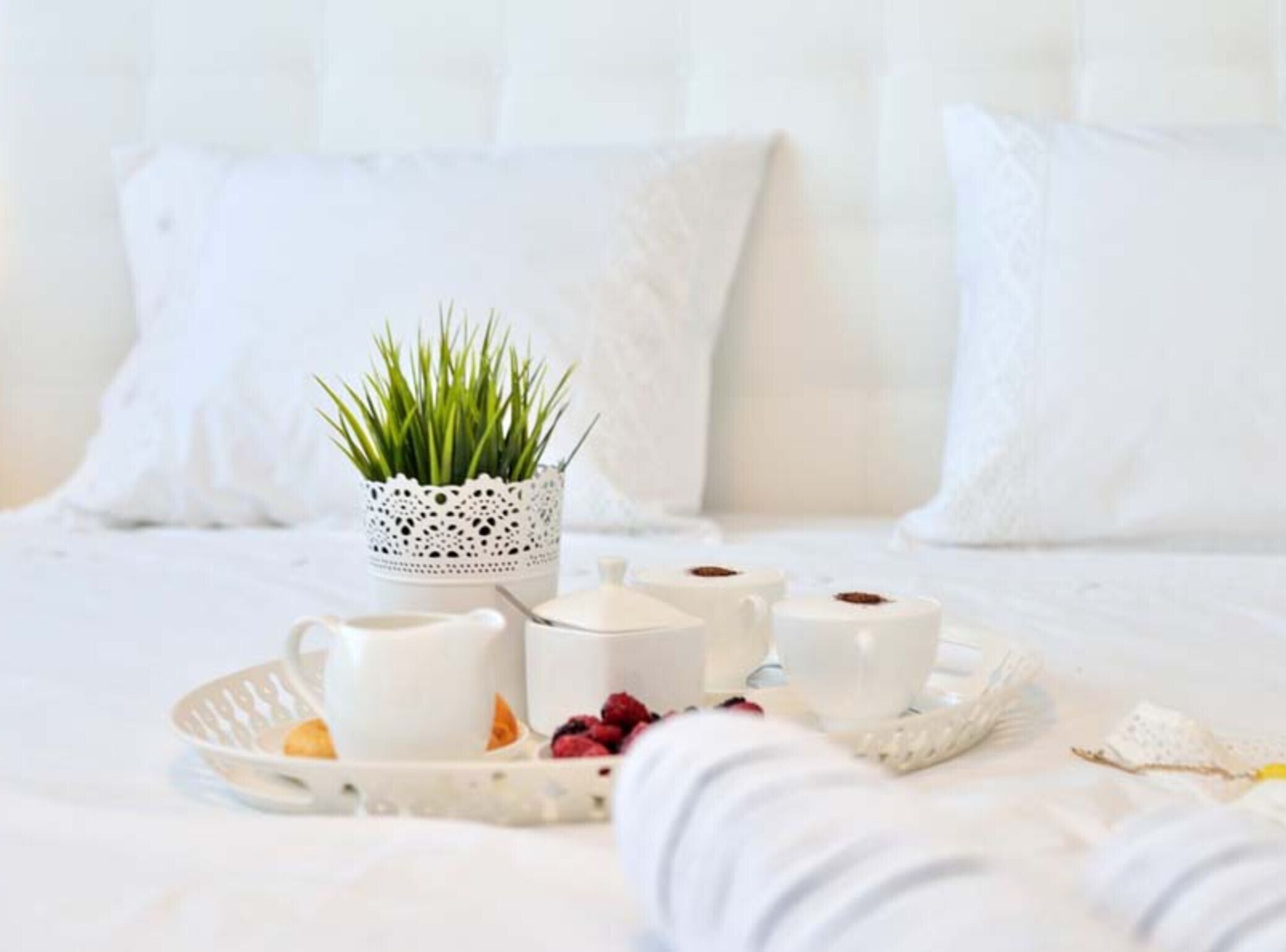 Ein ordentlich gedeckter Frühstückstisch mit weißer Karaffe, Tassen und einer kleinen Pflanze auf einem Bett mit weißer Bettwäsche und Kissen.