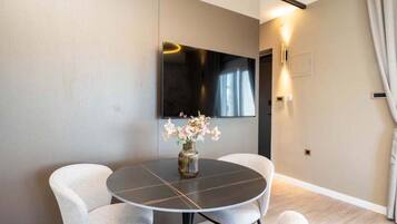 A modern room with a round table, two chairs, a vase with flowers, a flat-screen TV mounted on the wall, and a door with a light switch.