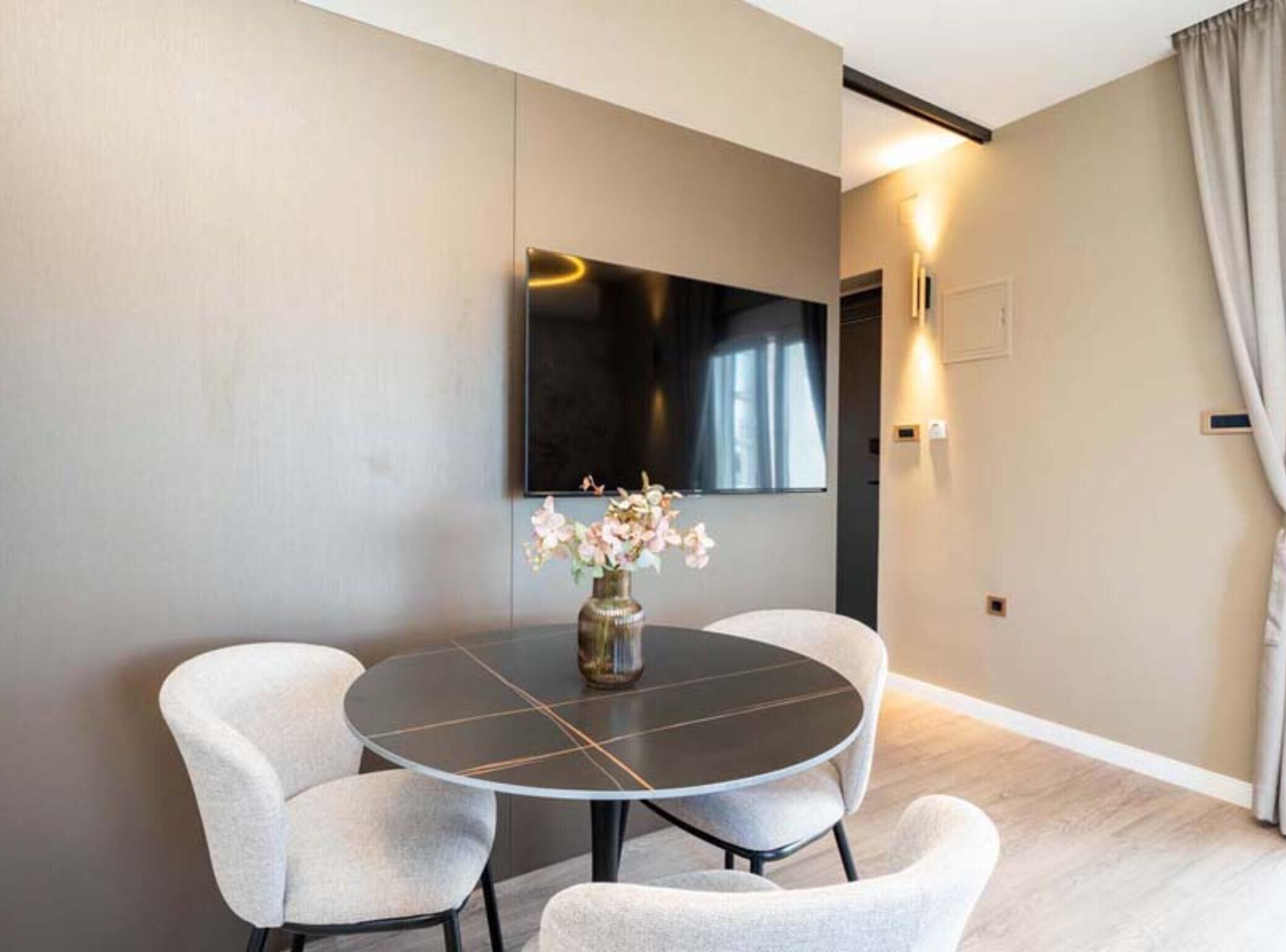 A modern room with a round table, two chairs, a vase with flowers, a flat-screen TV mounted on the wall, and a door with a light switch.