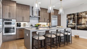 Una cocina moderna con electrodomésticos de acero inoxidable, una isla central con taburetes de bar, iluminación colgante y un cuadro abstracto de gran tamaño en la pared.
