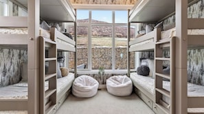 Un dormitorio tipo camarote con dos pufs, una ventana con vistas a un muro de piedra y árboles, y estanterías empotradas.