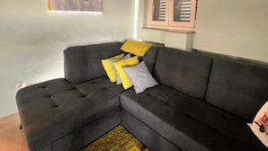 Ein dunkelgraues Ecksofa mit gelben und weißen Kissen, einem gelben Zierkissen und einem kleinen Teppich in einem Wohnzimmer mit Fenster und Tür im Hintergrund.