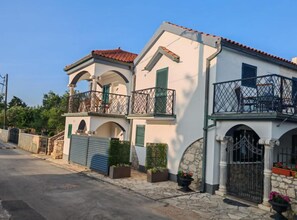 Un edificio de dos pisos con un balcón en el nivel superior, un muro de piedra y una puerta de metal.