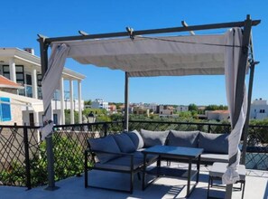Un espacio de estar techado al aire libre con sofá y mesa, una pérgola, y vistas a edificios en el fondo.