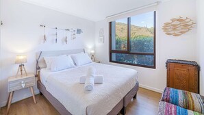 A neatly made bed with white linens and a rolled towel, a wooden nightstand with a lamp, a window with a view of a desert landscape, and a colorful woven bench.
