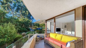 A balcony with a wicker sofa, glass railing, and a view of a mountain and greenery.