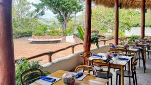 An outdoor restaurant with thatched roofs, wooden tables, and chairs, surrounded by greenery and a view of hills. - Casa Altavista 3 Bedroom 4 Bath Oceanview Home with 2 Pools, Patios, and Balconies (San Juan del Sur)