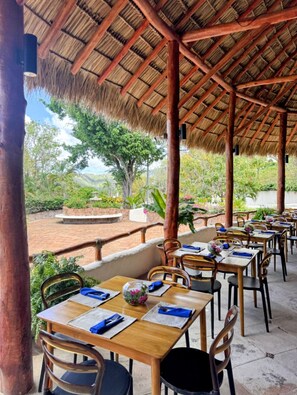 Een openluchtrestaurant met rieten daken, houten tafels en stoelen, omgeven door groen en uitzicht op heuvels.