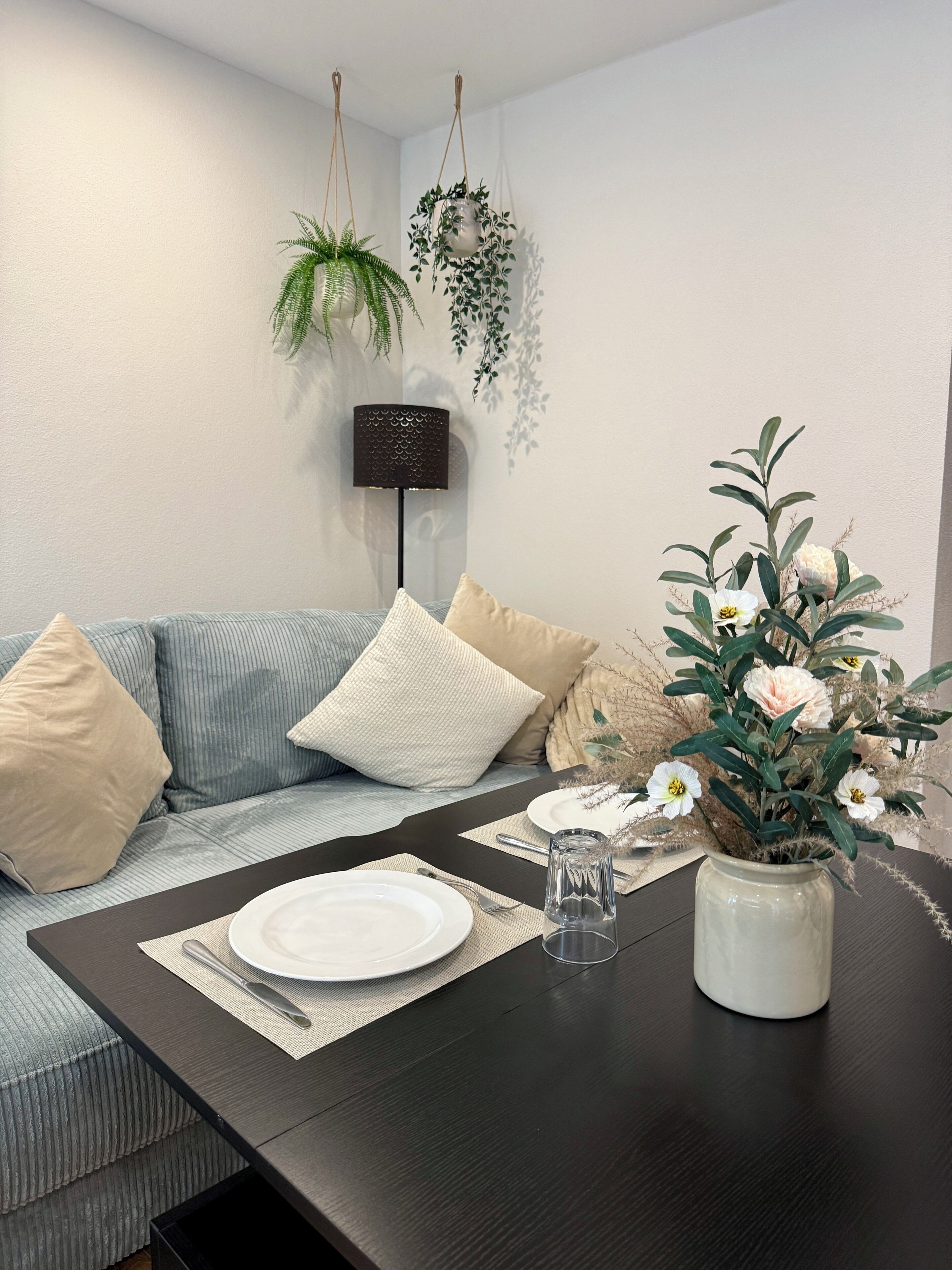 Un coin repas avec une table en bois foncé mise pour deux personnes, un canapé avec des coussins à rayures et une plante suspendue.