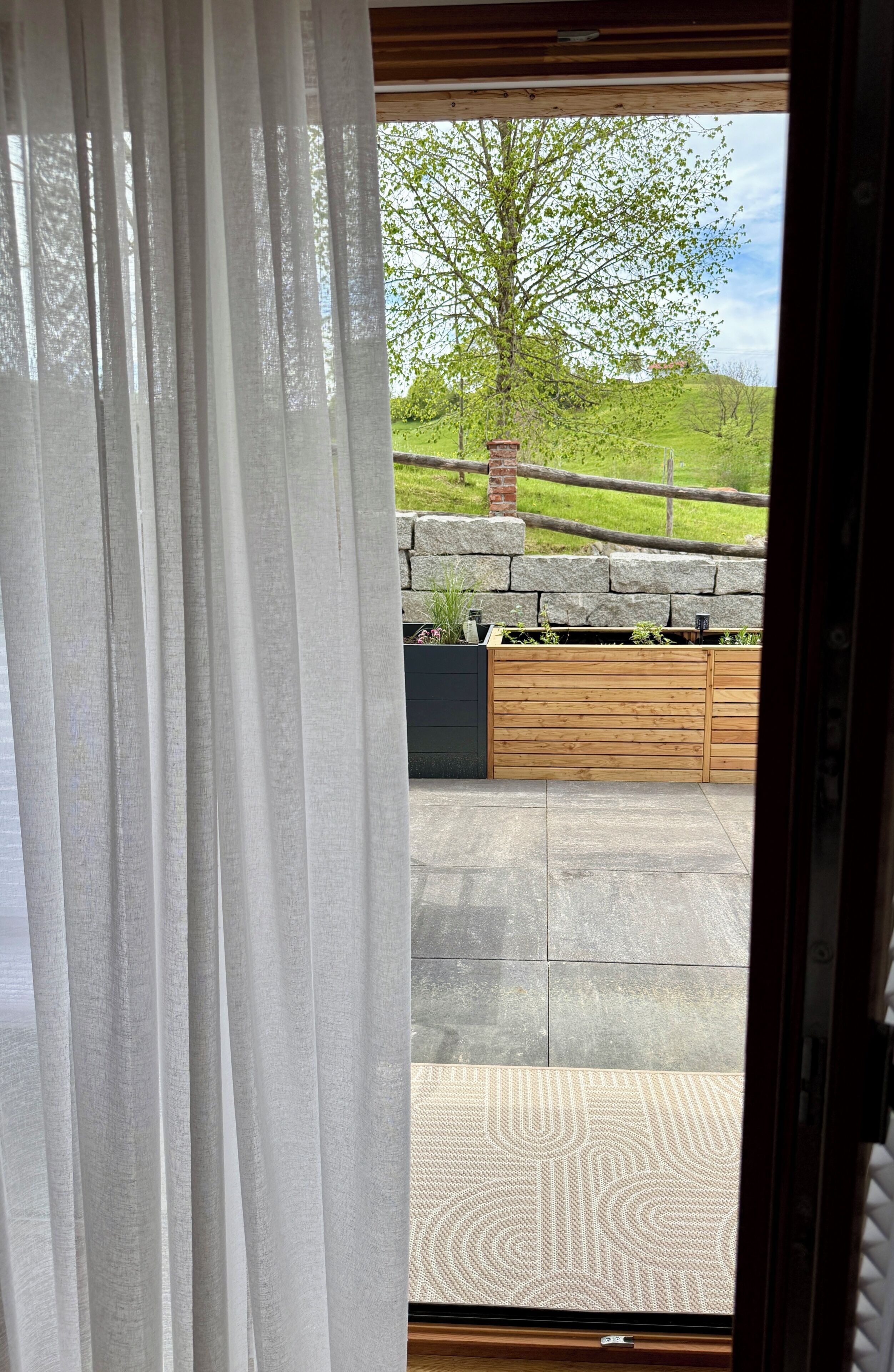 Blick durch ein Fenster mit Vorhang, eine Steinmauer, ein Holzzaun und ein grasbewachsener Hügel.