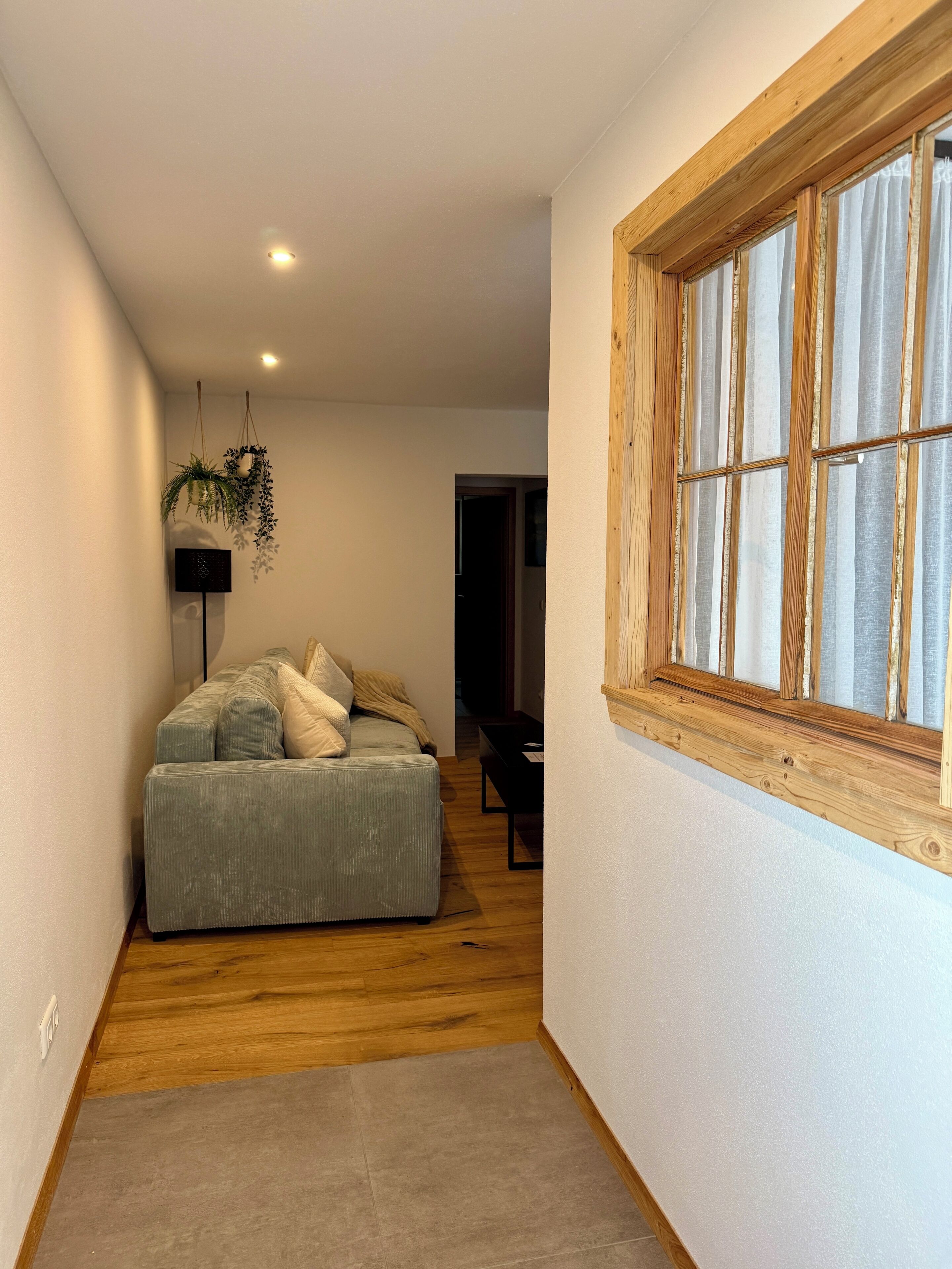A narrow hallway with a wooden floor, a grey sofa with cushions, a hanging plant, and a window with wooden framing.