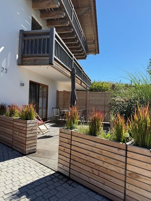 Ein zweistöckiges Gebäude mit einem Balkon, Außenbereich zum Sitzen und Holz-Pflanzgefäßen mit Pflanzen.