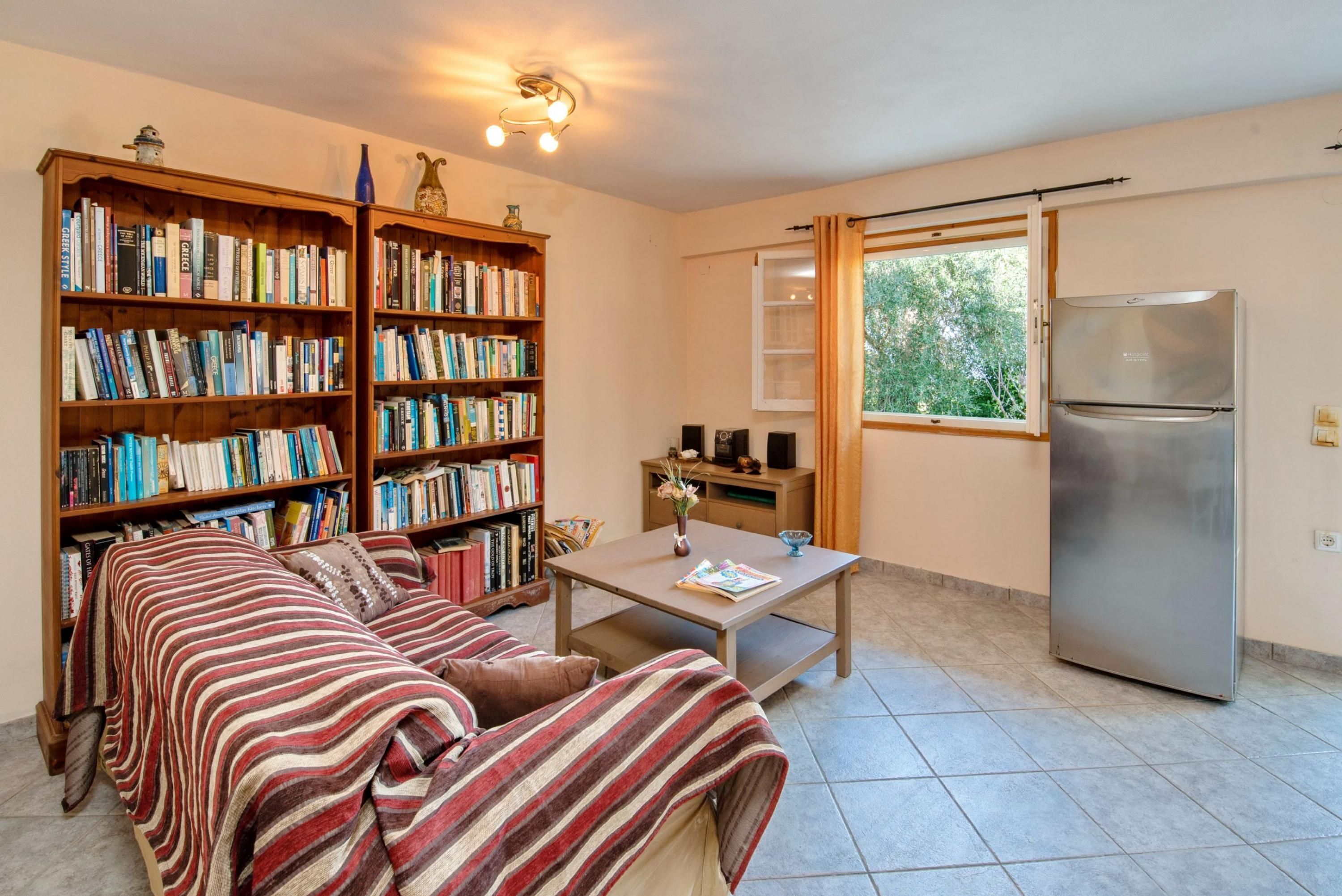A cozy living room with a sofa, two bookshelves filled with books, a coffee table with a vase and a book, a refrigerator, and a window with curtains.
