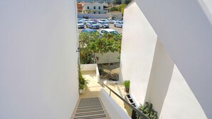 Vista de uma varanda com vista para um parque de estacionamento, com vários carros, uma escadaria e um edifício com fachada branca ao fundo.