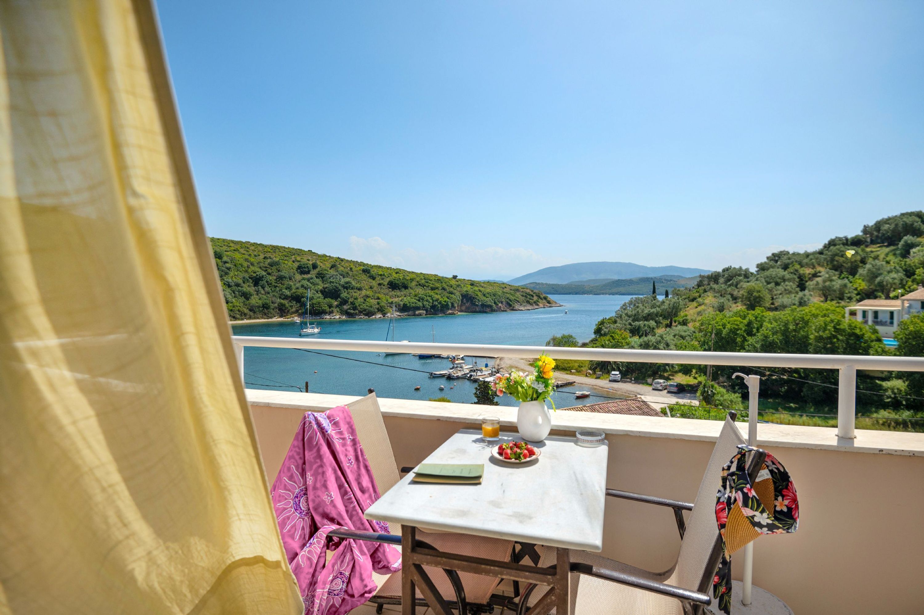 A balcony with a table, chairs, and a view of a lake with boats and hills.