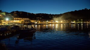 Un puerto deportivo en la costa al atardecer, con barcos amarrados, edificios al fondo y una colina con luces.