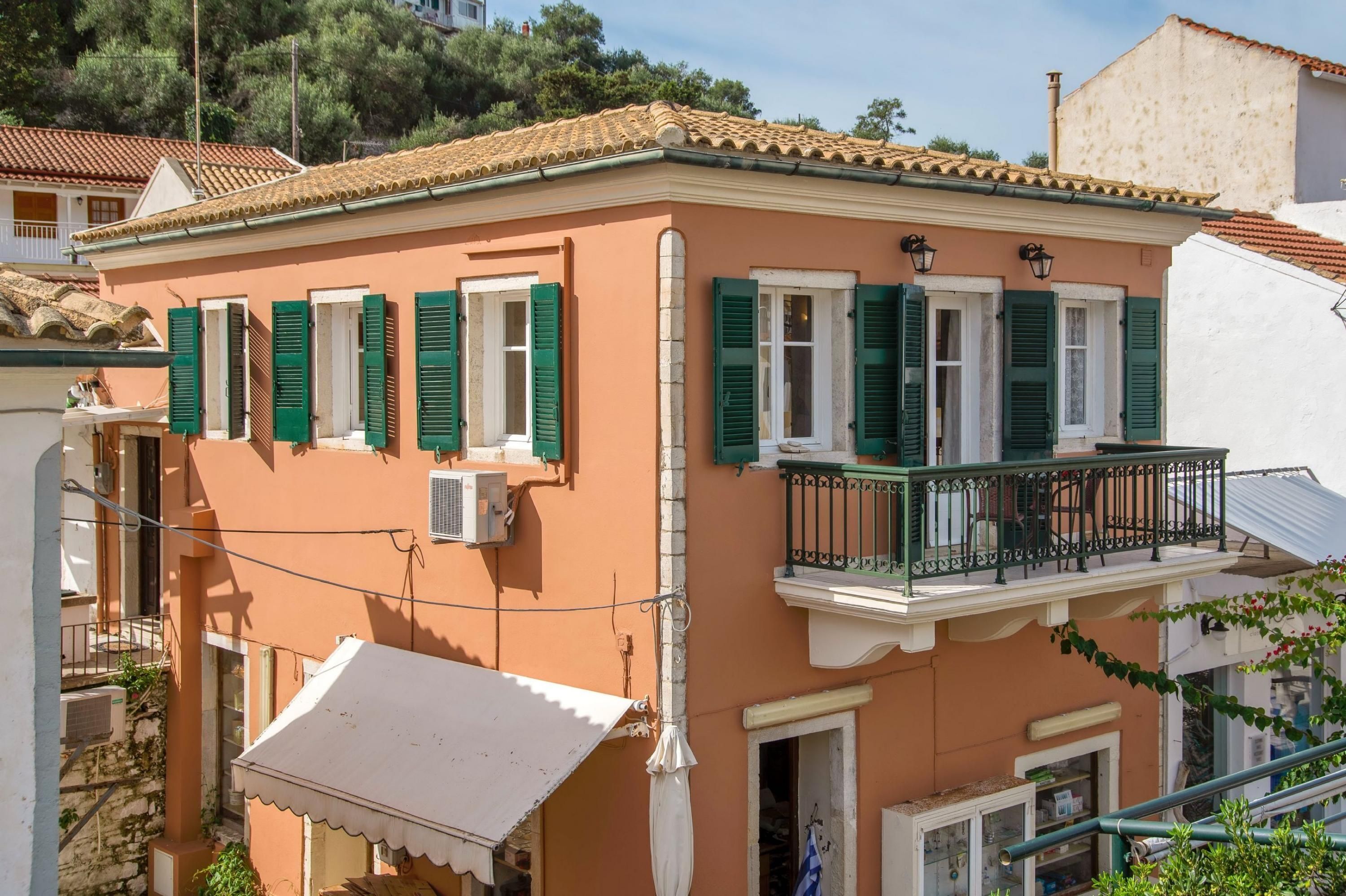 Ein zweigeschossiges Gebäude mit grünen Fensterläden, einem Balkon mit schwarzem Geländer und einem weißen Markisenvorbau.