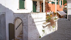 Une rue étroite bordée d’un immeuble blanc, caractérisé par un escalier en pierre et des volets verts.