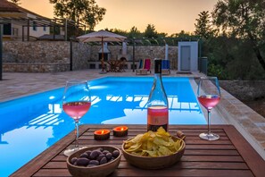 Une table au bord de la piscine, avec deux verres de vin, une bouteille de vin, des bols d’olives et des chips, et un mur de pierre en arrière-plan.