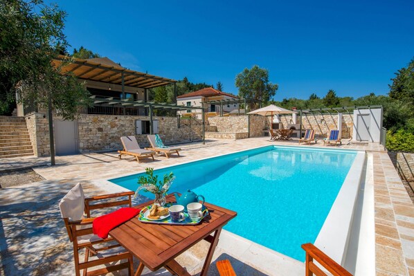 Une piscine entourée de chaises longues et d’une table dressée pour un repas, le tout bordé de murs en pierre et surmonté d’une pergola.